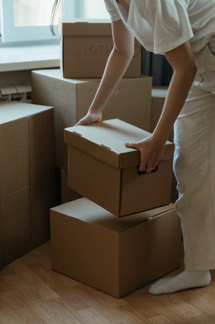 A person, dressed in light-colored clothing, is seen in a room with natural light from a nearby window, carefully lifting a cardboard box used for packing belongings during a home relocation. The individual's hands are positioned on the sides of the box, which is plain brown with no visible markings except for a faintly written label. Multiple similar boxes are stacked nearby, some placed directly on the wooden floor, while others are arranged on a piece of furniture or other boxes, indicating an organized packing process. The scene reflects typical packing activities associated with furniture transport and moving logistics managed by Hounslow Removals, including the safe handling of household items, the use of cardboard packaging for protection, and careful stacking to facilitate loading into a removal van. This visual captures the meticulous nature of packing and preparing items for a house removal, with attention to proper handling and organization essential for ensuring a smooth home relocation.