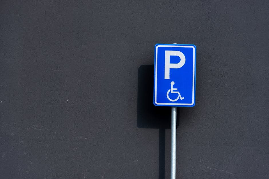 A close-up photograph of a blue parking sign with a white 'P' symbol and a wheelchair icon, indicating accessible parking, mounted on a metal pole attached to a dark grey exterior wall. The sign casts a shadow on the wall behind it. The environment appears to be outdoors, with the sign positioned on a paved area near the entrance of a property, suggesting access for mobility-impaired individuals during overall house removals or furniture transport activities. This image is relevant to house relocation logistics managed by Hounslow Removals, especially regarding parking permits for large removals at Highbury, as shown on the page titled 'Hounslow council parking permits for large removals, Highbury'.