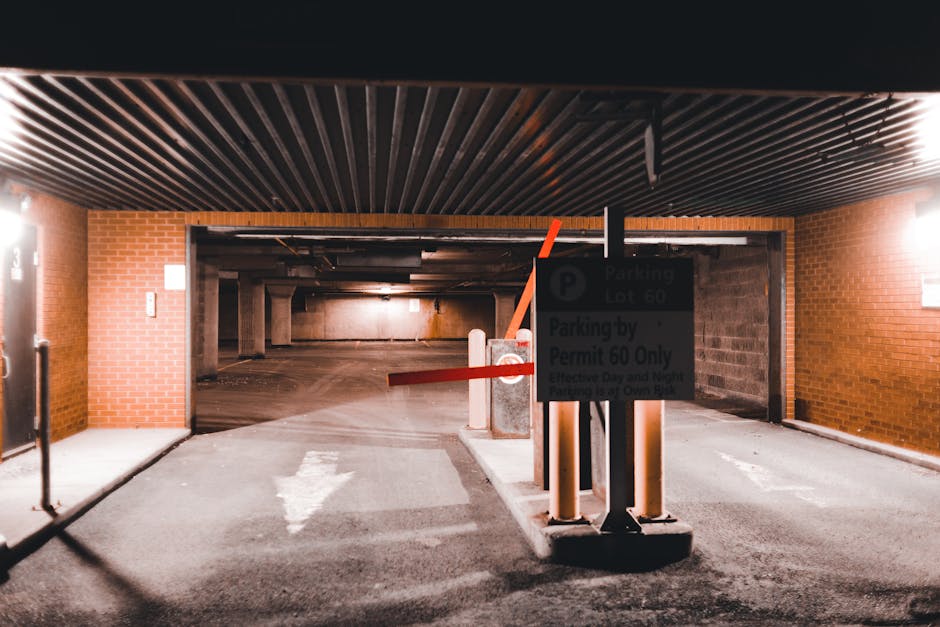 View of an underground parking garage entrance with a partially raised red barrier arm and a sign indicating parking restrictions for large removals, situated on a paved driveway leading to an interior parking area. The garage features brick walls on either side, a metal slatted ceiling, and concrete supports. Inside the parking lot, there are concrete pillars and low ceiling sections, suggesting a multi-level structure. The setting appears to be prepared for home relocation or furniture transport, with the barrier controlling access and supporting parking permits for large move vehicles, as managed by Hounslow Removals. The garage is illuminated with artificial lighting, and the overall scene captures the logistical environment involved in professional removals and packing services, aligning with house moving processes.