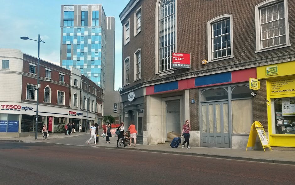View of a busy high street in Hounslow with pedestrians walking along the sidewalk, some carrying shopping bags or pulling wheeled suitcases, in front of a row of commercial buildings. On the left, a Tesco Express convenience store is visible with its logo and storefront, while on the right, a property with a 'To Let' sign and a yellow newsagent shop with various notices in its window. In the foreground, a woman wearing sunglasses and a pink top pulls a suitcase as she walks past the shop, while other pedestrians are scattered across the sidewalk. The background features a mix of older brick buildings and modern high-rise structures, with a streetlamp and a clear sky above. This scene exemplifies a typical urban environment where house removals or furniture transport might take place, involving packaging, loading, and logistical planning supported by companies like Hounslow Removals.