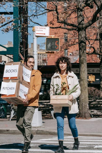 Two individuals walking across a street during daylight, each carrying packed cardboard boxes labeled 'BOOKS' and 'KITCHEN'. The man on the left has dark hair and a beard, wearing a brown jacket and grey pants, while holding a large box with multiple smaller boxes stacked inside. The woman on the right has curly dark hair, wearing a cream-colored coat, a patterned yellow top, and blue jeans, carrying a wooden crate with green leafy plants. Behind them is a traffic light showing a red signal, a digital countdown timer displaying five seconds, and a tree with bare branches against a background of brick buildings and shop signs, indicating an urban environment suitable for home relocation or furniture transport. This scene illustrates packing and moving activities, consistent with services offered by Hounslow Removals for local house moves, emphasizing loading, packing, and moving logistics in a city setting.