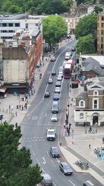An aerial view of a busy urban street during daytime, showing multiple vehicles including cars, vans, and a double-decker bus traveling along the road. On the left, a building with a sign reading 'La Pirates' is visible, alongside several pedestrians walking on the sidewalk. The street is lined with trees providing greenery amidst the urban environment. On the right, there is a small white building with a pitched roof and a bicycle parked nearby, alongside a few more pedestrians and cyclists. The area appears to be part of a city neighbourhood, with residential and commercial structures. This scene represents typical city traffic and activity, which might be relevant in the context of house relocations or moving services, such as those offered by Hounslow Removals, especially for same-day last-minute home relocation or furniture transport in Hounslow.