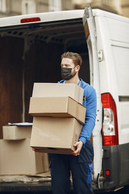A professional mover from Hounslow Removals is inside a bright, spacious room with natural daylight coming through large, arched windows. He is wearing a navy blue uniform with the company's logo on the chest and black work shoes. The mover is in the process of packing or preparing for furniture transport, holding a cardboard box sealed with red tape in his right hand, while two other boxes, also sealed, are placed on the wooden floor in front of him. The room has plain white walls and a light-colored wooden floor, with no furniture visible. The scene captures the typical packing and loading process involved in home relocation, with the mover ready to load the boxes into a van for efficient moving services as part of the comprehensive removals offered by Hounslow Removals for same-day, last-minute house moves in Highbury.