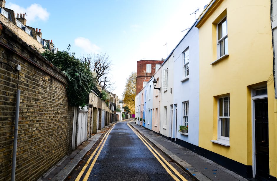 A narrow, slightly curved street in Hounslow during daytime, with a wet asphalt road marked by double yellow lines along the curb. On the left, an aged brick wall with climbing greenery separates the street from residential properties, which have small front gardens and varied door designs. On the right, a row of terraced houses features a mix of pastel-colored facades including white, yellow, and light blue, with white-framed sash windows, some with flower boxes. The ground-floor windows of the houses are aligned with the pavement, which is paved with grey concrete slabs. In the background, a red-brick building with multiple stories rises above the houses, partially obscured by leafless trees indicating autumn or winter. Light from the overcast sky illuminates the scene evenly, highlighting the textures of the buildings and street. Hounslow Removals services are involved in facilitating home relocation in such confined urban environments, where careful furniture transport and packing are necessary due to limited street space and narrow passages.