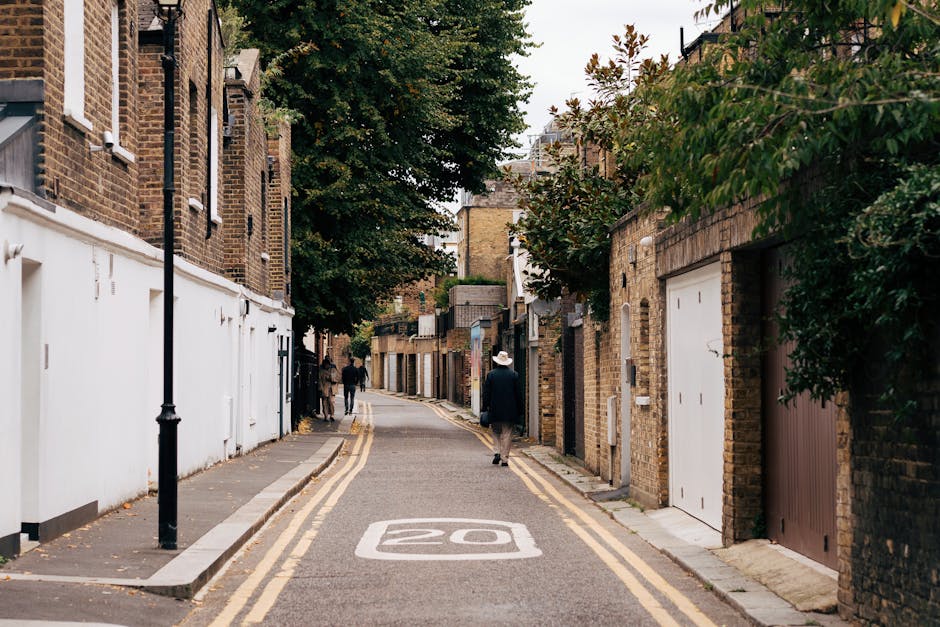 A narrow residential street in Hounslow lined with brick houses and small garages, with a road marked with a 20 mph speed limit. Several individuals are visible in the distance, including a person wearing a white wide-brimmed hat and dark clothing, walking away from the camera. The pavement is narrow, with black lamp posts along one side, and there are trees with green foliage extending over the buildings and street. The scene illustrates the challenges residential house removals and furniture transport face in tight urban streets, requiring careful planning by moving companies such as Hounslow Removals. Items such as cardboard boxes, packed furniture wrapped in protective materials, and moving equipment may be involved in the process of packing and loading for home relocation, especially navigating restricted space and dealing with uneven or limited access points typical of such streets.