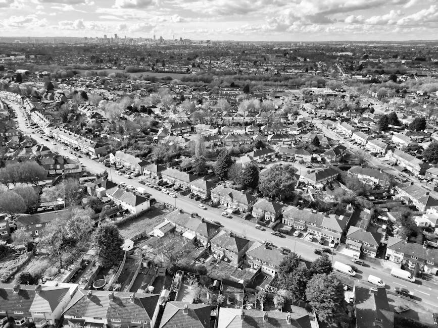 An aerial view of a busy urban street during daytime, with multiple lanes of moving traffic including cars, vans, and buses, lined by both modern and older brick buildings. Pedestrians are visible on