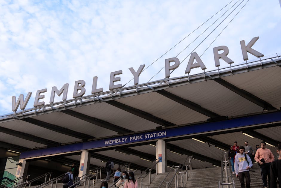 Exterior view of Wembley Park Station entrance with large, white, three-dimensional letters spelling out 'WEMBLEY PARK' positioned horizontally on the station's roof. The station signage indicating 'WEMBLEY PARK STATION' is mounted below the roof on a blue strip. Several commuters, some wearing face masks and carrying backpacks, are seen descending the stairs outside the station on an overcast day with a partly cloudy sky. The station structure includes metal railings and steps leading up from the pavement, with visible electrical wiring overhead. This scene captures the typical busy atmosphere of a London transport hub, relevant for home relocation and moving services offered by Hounslow Removals, as part of packing and furniture transport processes near Hounslow West station.