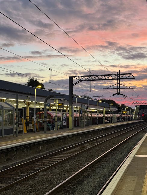 View of Hounslow West railway station at dusk with multiple train carriages parked on the platforms, illuminated by overhead lighting. The station features metal canopy structures over the waiting areas, with signage visible behind the fencing. Overhead electric wires extend across the sky, which displays a sunset with pink, orange, and purple hues. The train tracks run parallel to the platform, bordered by concrete paving and gravel beds. In the background, residential buildings and trees are faintly visible. This scene illustrates a typical urban train station environment, where Hounslow Removals may assist with home relocation or furniture transport involving station-to-station transfers, packing, and loading processes.
