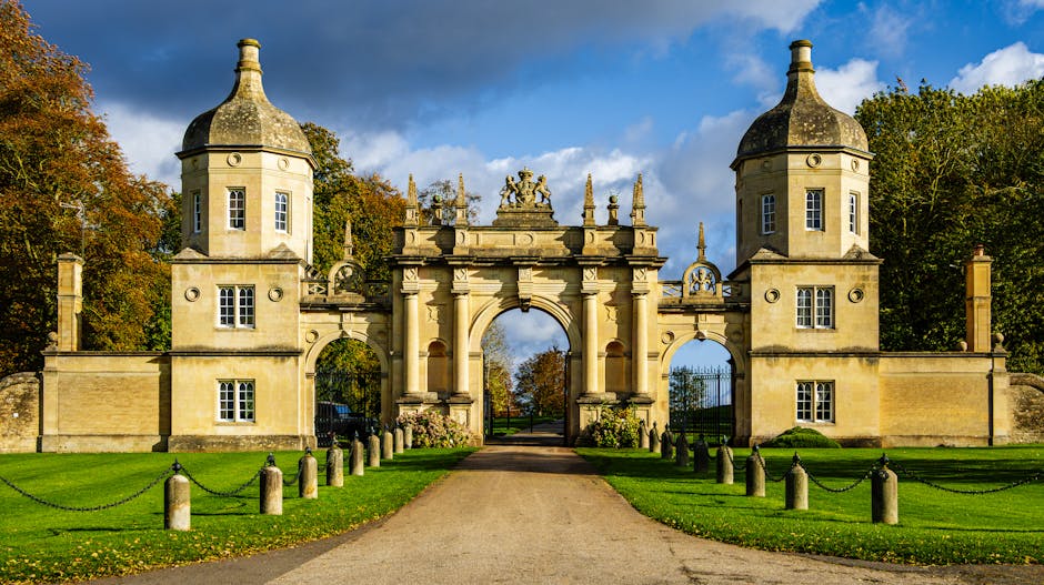 A historic stone archway with two rounded towers on either side, featuring conical roofs, situated at the entrance to Syon Park. The archway has decorative statues on top and intricate carvings, with a large central opening leading to a pathway lined with low stone bollards connected by chains, guiding visitors through the park. The surrounding area includes well-maintained green lawns and mature trees with autumn foliage, under a partly cloudy sky. This scene illustrates the type of location where house removals or furniture transport services by Hounslow Removals might facilitate access for home relocation or loading of bulky items, with the archway serving as a notable landmark in the process of delivering or collecting large objects during a move.