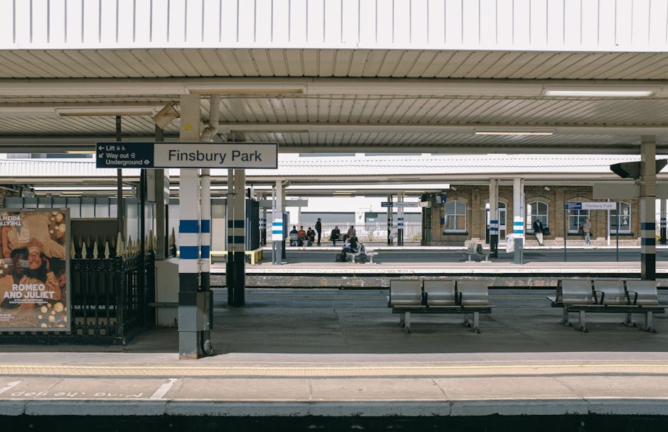 The image shows Finsbury Park train station platform during daytime with several passengers seated on benches and walking along the platform. The station signage is visible, indicating the name 'Finsbury Park' and directions for lifts and underground access. The platform is covered by a roof with fluorescent lighting, and the flooring is made of concrete with distinct safety markings. In the foreground, there are a few empty benches, and along the edge of the platform, a black metal fence separates passengers from the train tracks. The background features additional platforms, station buildings with brick facades, and some pedestrians waiting or walking. This scene captures a typical busy railway station environment, relevant to house removals and moving logistics where efficient loading and transport of furniture and belongings are integral, with the station serving as a key transit point for relocations managed by Hounslow Removals.
