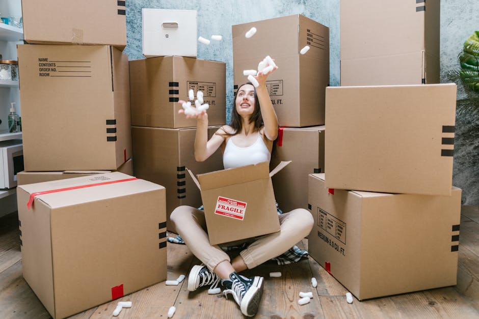 A young woman with long dark hair sitting on a wooden floor inside a room, surrounded by numerous large cardboard moving boxes, some sealed with red tape and others open. She is wearing a white sleeveless top, beige pants, and black sneakers with white soles. She is smiling and tossing foam packing peanuts into the air, with additional packing materials scattered around her. The boxes are stacked in various heights against a textured blue and gray wall, indicating an active home relocation process. A small part of a white kitchen shelf and a green potted plant are visible in the background, suggesting the setting is a residential space. The scene captures the packing and moving stage of a house removal, with [COMPANY_NAME] involved in the logistics and transport of furnishings and belongings, as highlighted on the [PAGE_TITLE] page of hounslowremovals.org.uk.